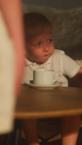niño aburrido se siente inquieto en la silla en la mesa. niño pequeño cansado de la fiesta de té sin interés con la hermana en casa. niño travieso se inclina para mostrar con tazas en la habitación