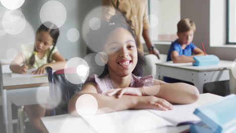 animation of white bokeh light spots over smiling biracial girl at desk in diverse class