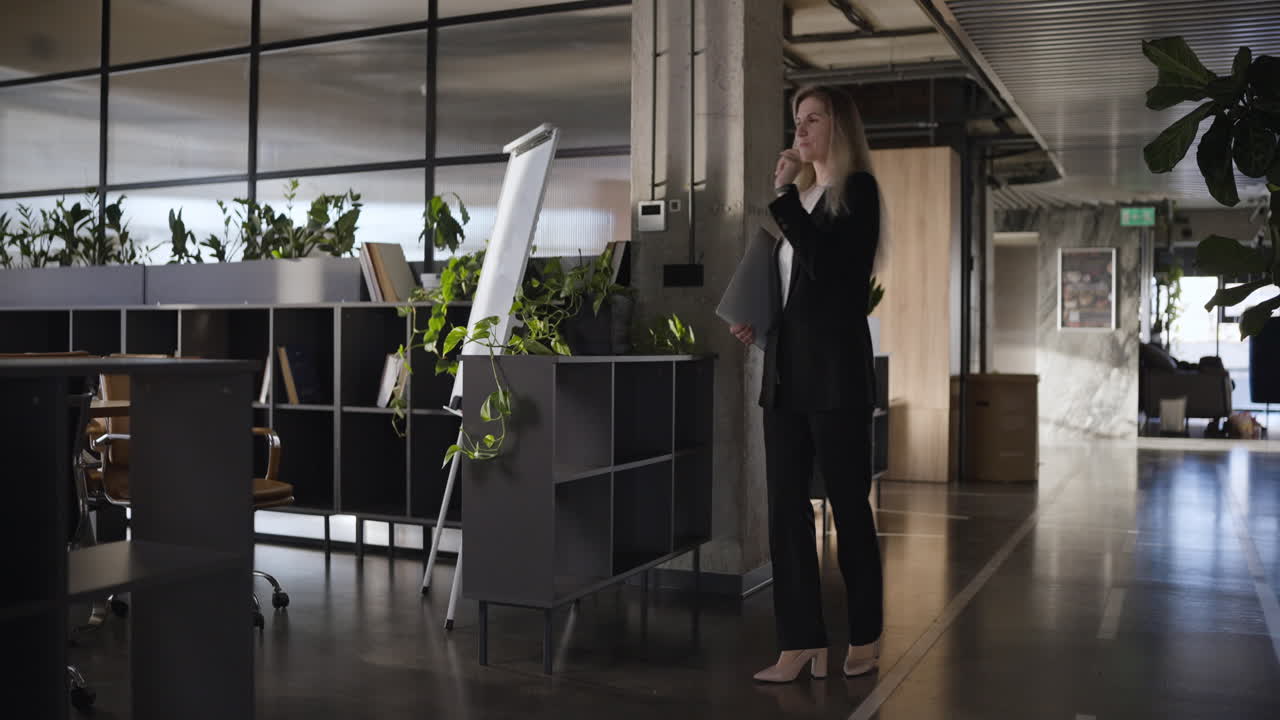 Woman in business suit walking in a modern office, carrying laptop and documents