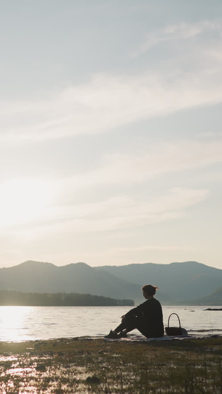 여성은 피크닉에서 물에 돌을 던집니다. 음식 바구니를 가진 여성 실루은 따뜻한 가을 저녁에 자연의 아름다움에 둘러싸인 평화로운 피크닉을 혼자 즐기고 있습니다.