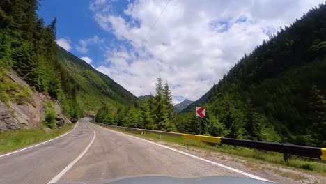 Driving-on-Transfagarasan-Mountain-Road,-Surrounded-by-Tall-and-Green-Fir-Trees-with-a-Clear-Blue-Sky-and-Thick-White-Clouds-in-Romania