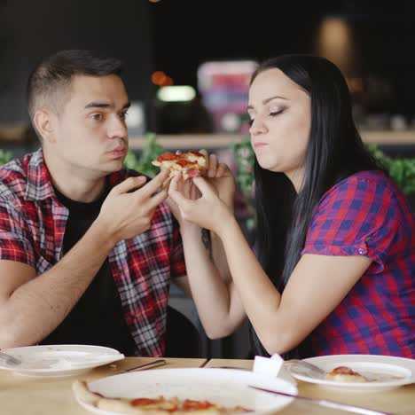romantic couple are eating one slice of pizza together in the pizzeria on the background of decorative plants