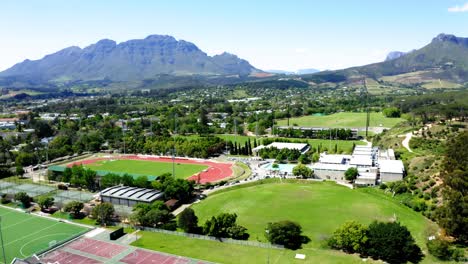 aerial drone rotating, university college sports athletics stadium, track and field, tennis courts, olympic swimming pool, mountains and trees in background, stellenbosch, coetzenburg