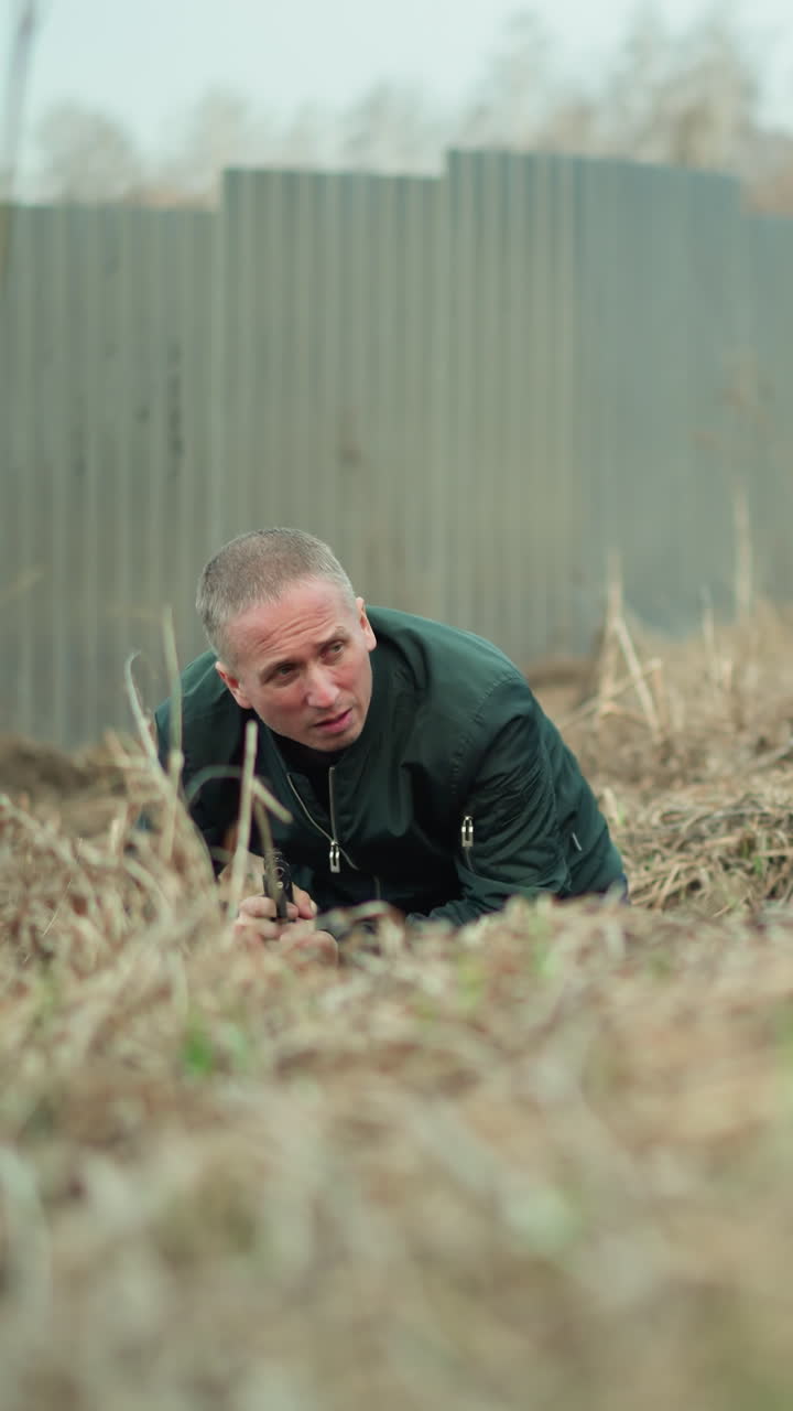 una vista cercana de un hombre con una chaqueta verde arrastrándose a través de la hierba seca, sosteniendo una pistola apuntando a algo, situado cerca de una valla de metal corrugado en un entorno al aire libre