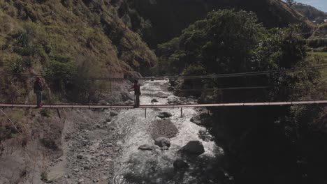 two men walking across a metal wire suspension bridge over rocky mountain water river in philippines stationary aerial medium wide
