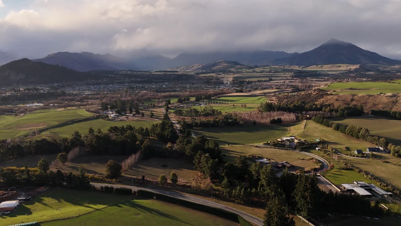 Aerial drone view flying backwards over green farmland near Wanaka. The shot reveals rural estates, the town, and the majestic Southern Alps under a moody sky in New Zealand South Island