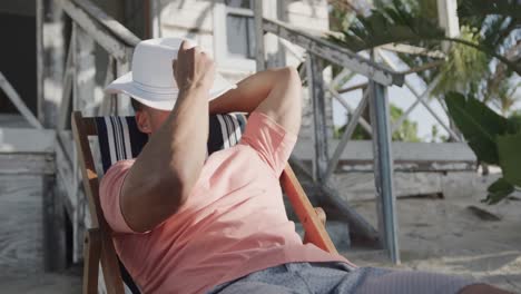 Entspannter-Gemischtrassiger-Mann-Sitzt-In-Der-Sonne-Mit-Hut-über-Dem-Gesicht-Vor-Dem-Strandhaus,-In-Zeitlupe