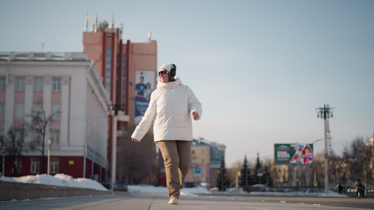 Free spirit walks along tiled pavement in snowy urban park swaying arms to rhythm while enjoying winter music in puffy white coat, beanie, sunglasses, headphones with carefree joyful vibe
