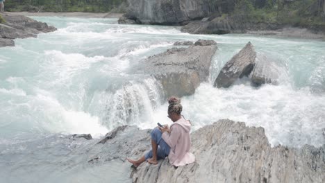 black lady on her phone sitting on the rocks near the edge of a waterfall (natural bridge lower falls) and beautiful scenic blue river in yoho national park