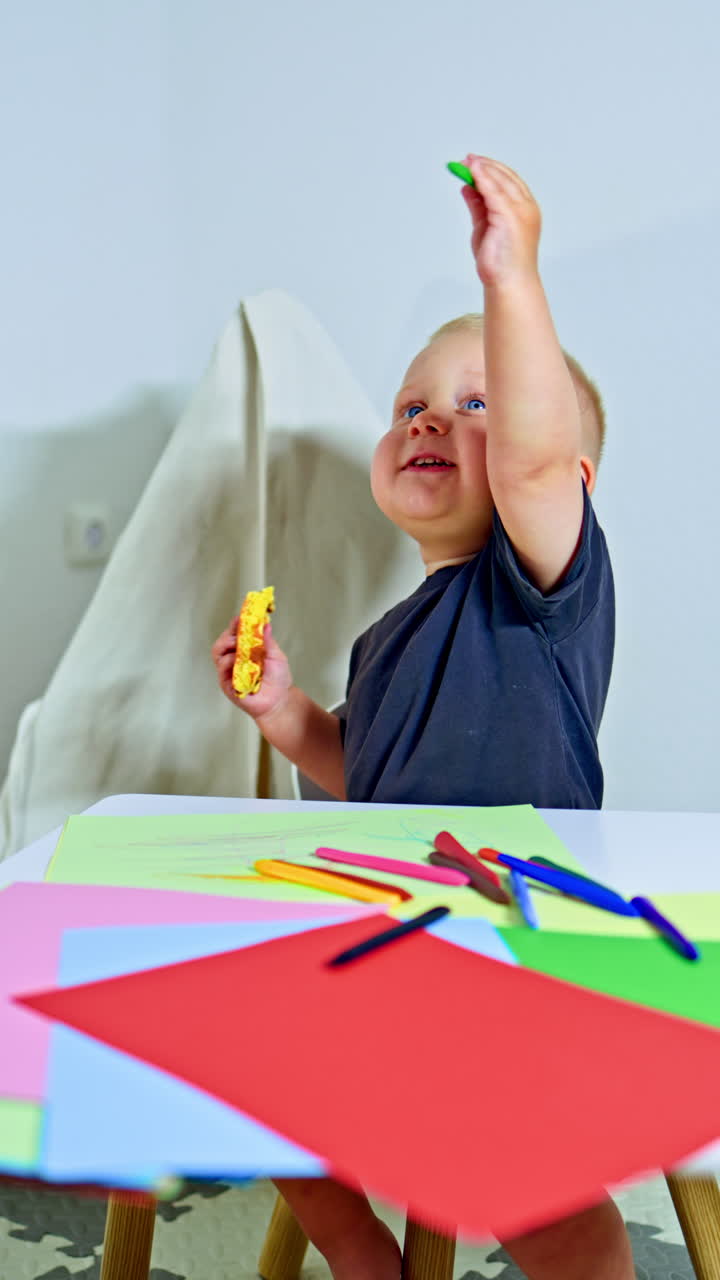 Cute toddler in a T-shirt sits at home at a children's table and draws with pencils. Child learns to draw on paper Child development while drawing with colored pencils. First pencils for toddlers