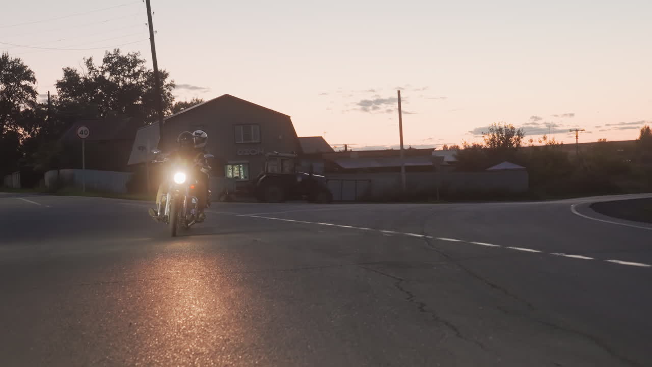 Motorbike riders journey through winding rural road at dusk, headlamp beaming brightly into camera, silhouetted by glowing sunset sky and scattered clouds, casting long shadows on smooth asphalt
