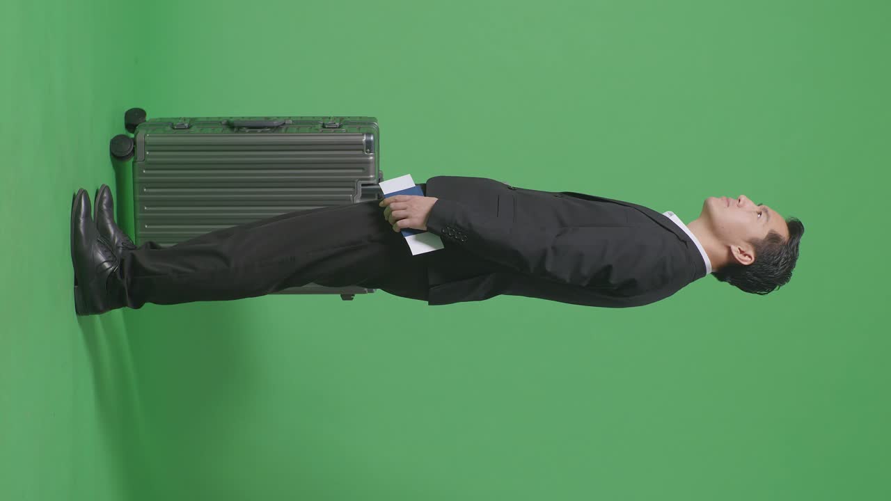 푸른 스크린 배경 스튜디오에 서있는 동안 여권을 들고 주변을 둘러보는 짐을 가진 아시아 사업가 여행자의 전체 측면 뷰
