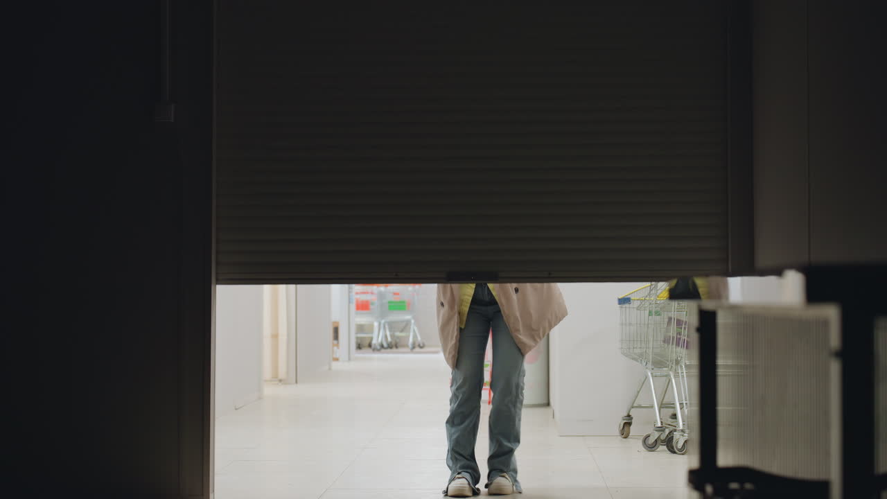 Laundry owner stands in corridor reaching upward as she jumps to pull down rolling steel door of laundry shop, wearing jacket and jeans, bright indoor setting with ceiling lights