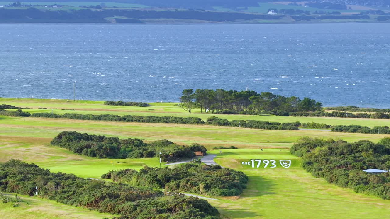 Wide shot pans over lush seaside golf course, vibrant greenery, and blue water under daylight