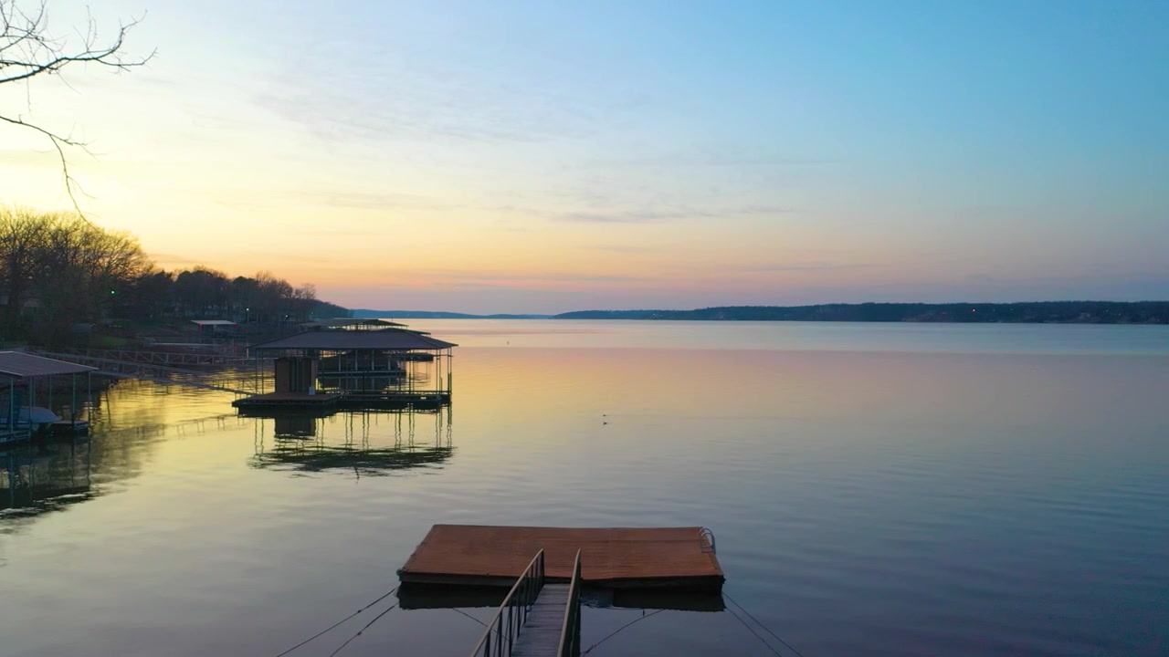 Wide aerial footage of a quiet reservoir neighborhood, with tree-lined shore, waterfront properties, covered docks, calm open water, and soft sunset colors fading across the horizon below.