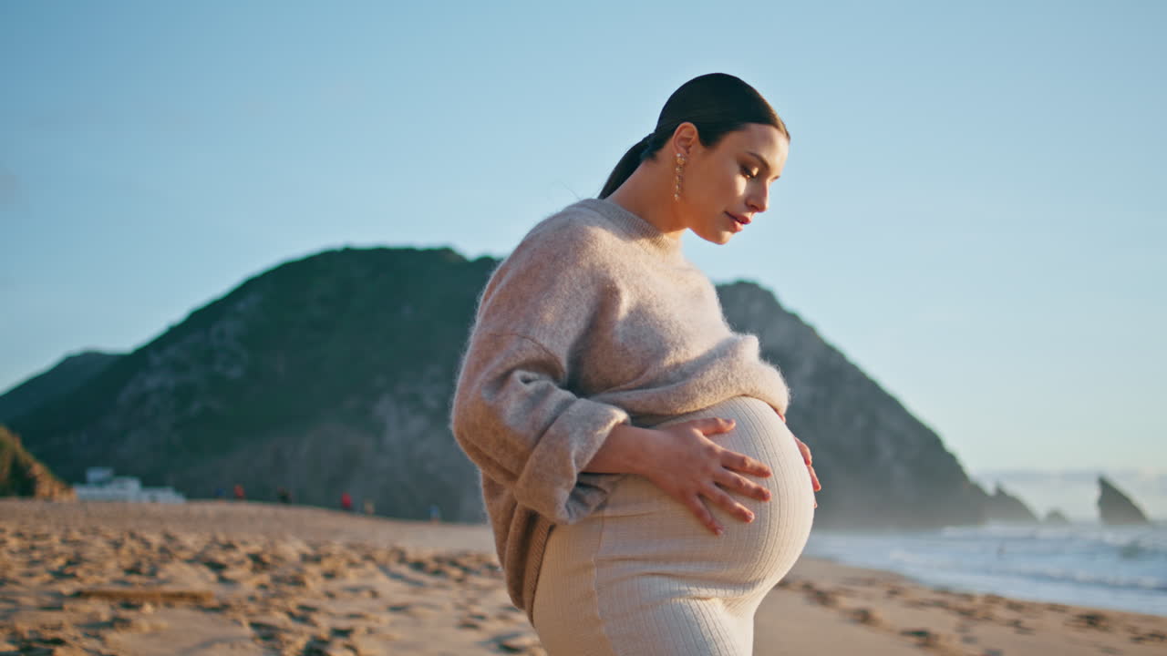 임신한 배를 만지는 여성이 해변에 서있는 은 저녁에 가까이 다가옵니다.