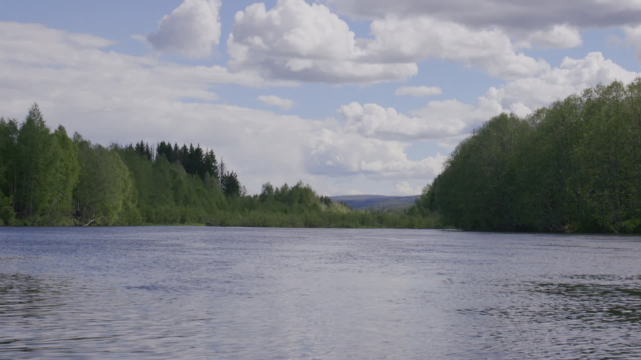 Wide view of calm river flowing between dense green forest under a cloudy blue sky with distant hills on the horizon. Filmed at Glomma, Norway's biggest river.
