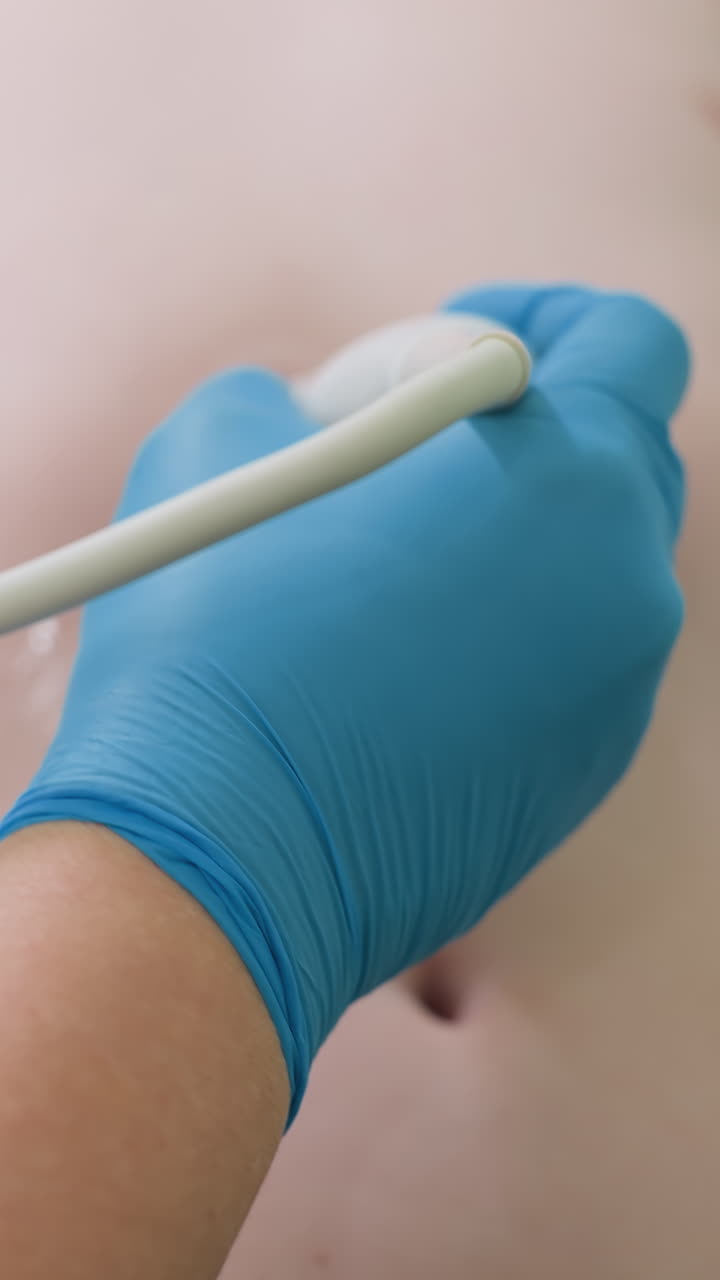 Manos enguantadas de azul deslizando una sonda sobre el pecho con una capa de gel, ambiente de valoración junto a la cama, presión suave y trazo firme, cable del dispositivo portátil visible, enfoque del médico en la calidad de la imagen.