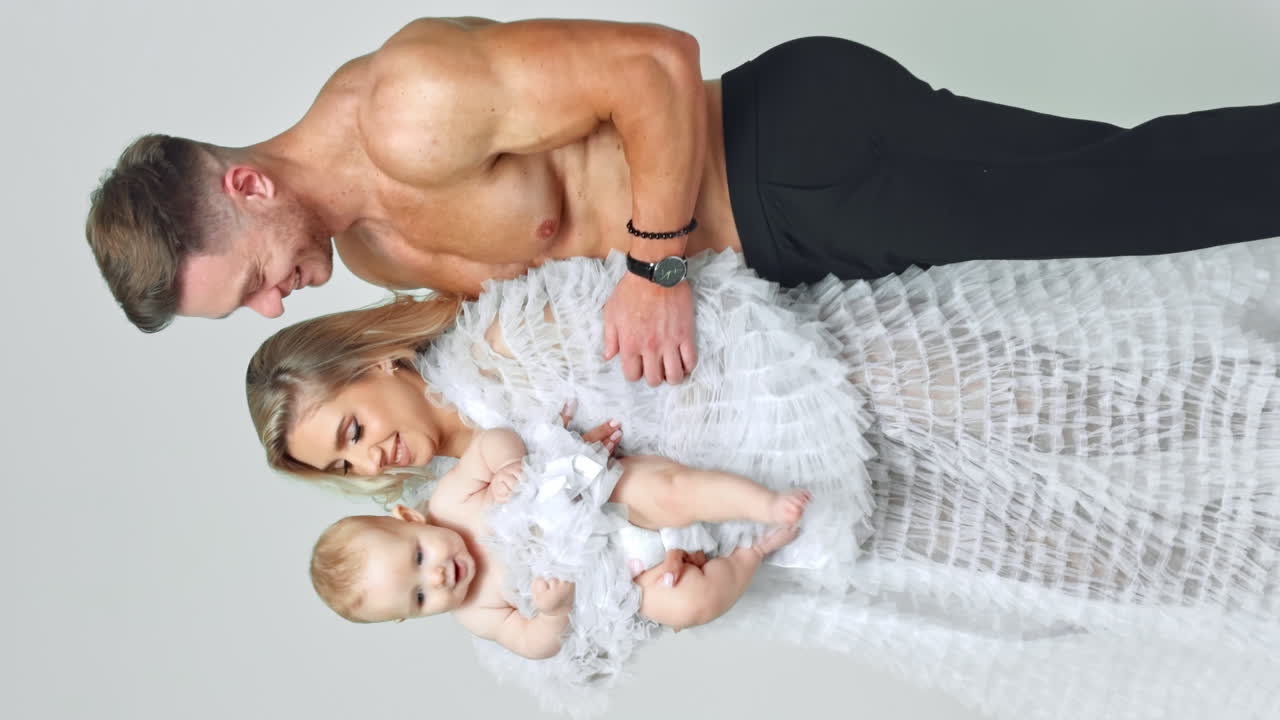 Smiling woman dancing with her child in hands. Muscular man stands behind holding a baby by the foot. White backdrop. Vertical screen.