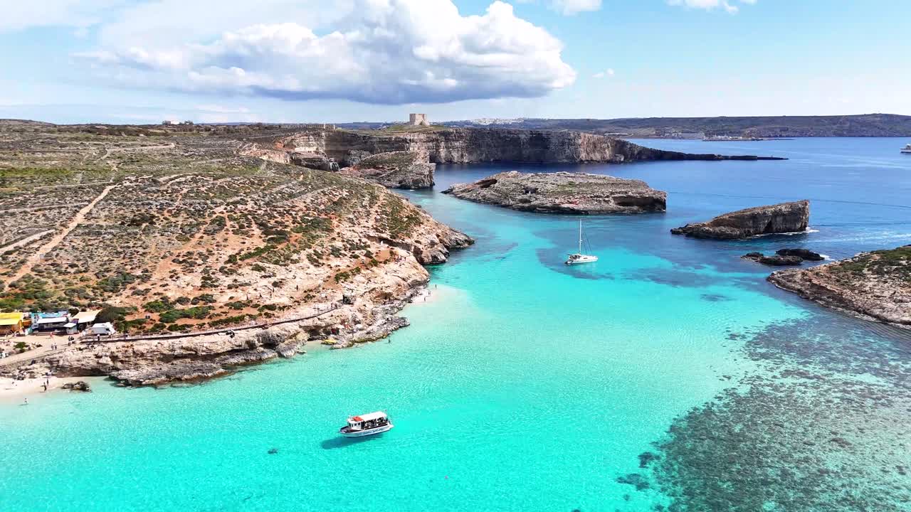 Veduta aerea della Laguna Blu a Comino, Malta, con acqua turchese, costa rocciosa, piccole imbarcazioni ancorate e un'ampia baia costiera di giorno.