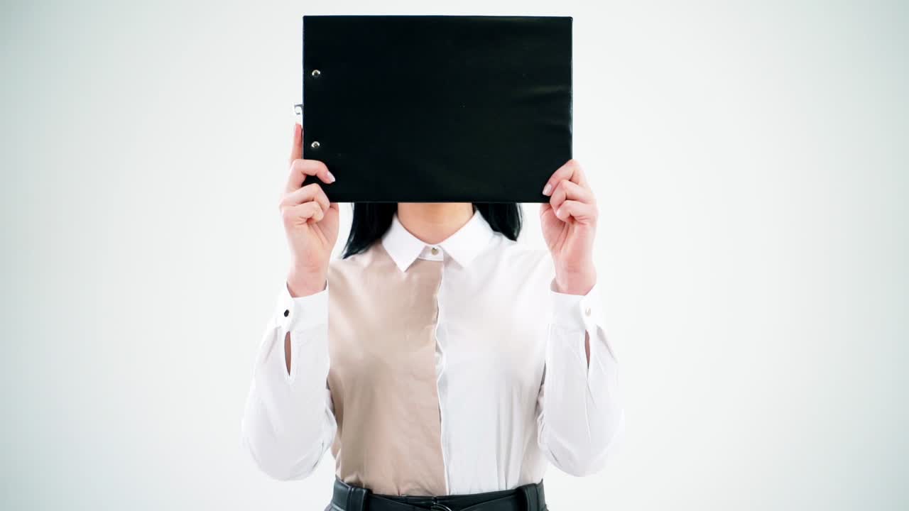 Black folder hides woman face. Attractive secretary opens her face hidden with a folder and smiles isolated on white wall. Businesswoman with positive emotions.