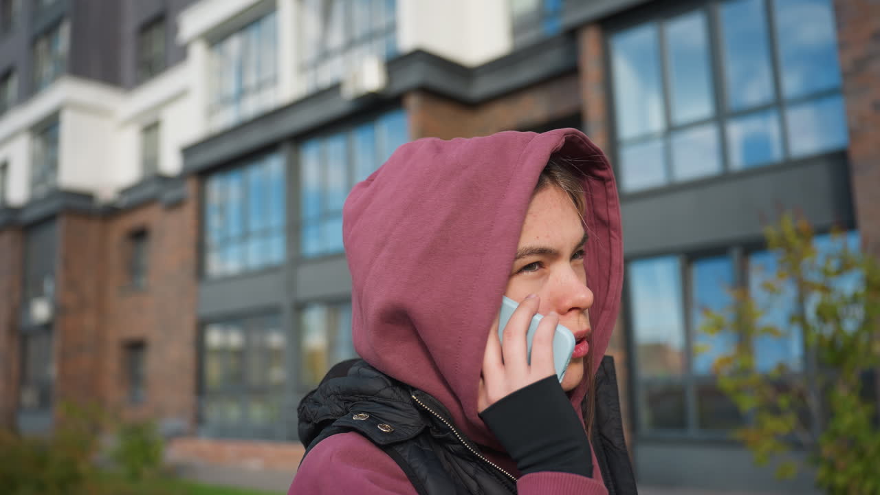 Student in mauve hoodie and black vest jacket speaks on phone with gentle confident smile under bright sunny sky in urban courtyard lined with modern high rise buildings and casual style