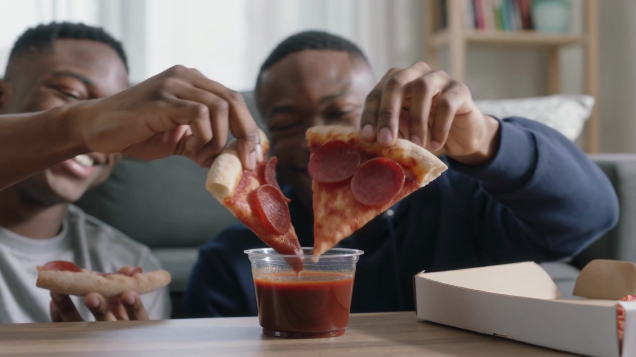 Holding slices, two men dipping pizza into red sauce cup on living room table, sharing snack