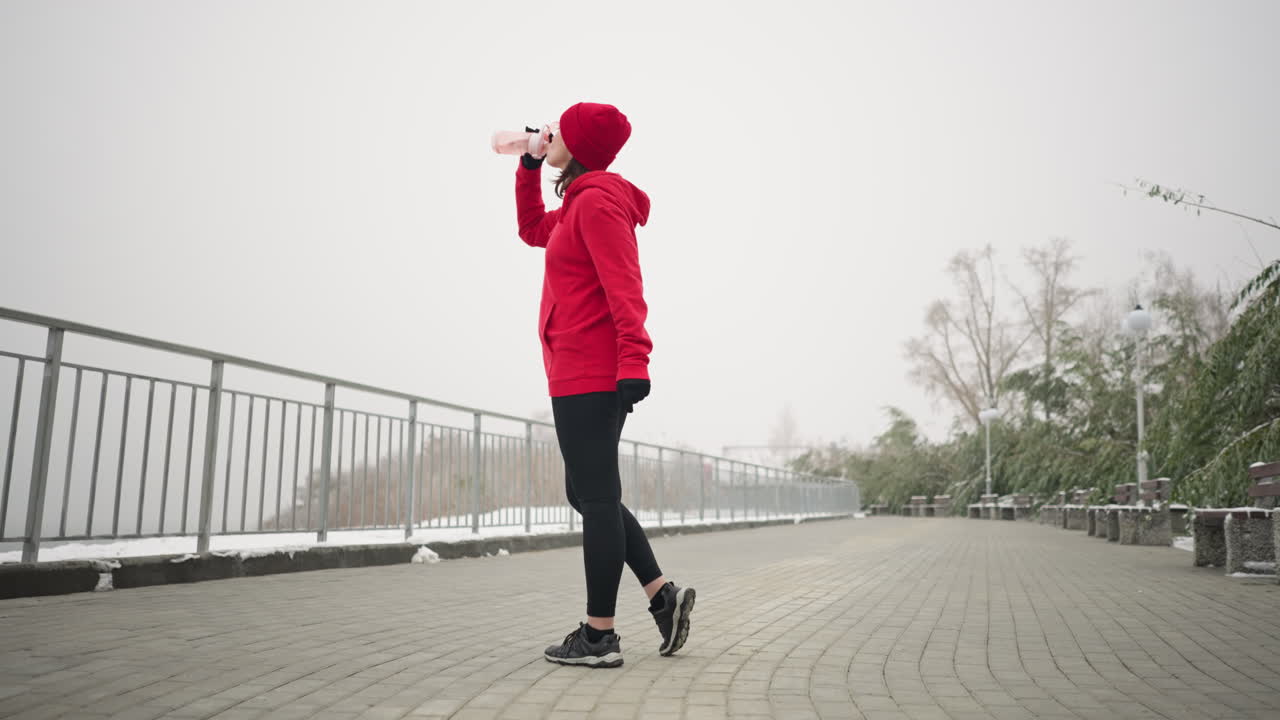 빨간 후디와 베니를 입은 여자가 은 겨울 공원 환경에서 핑크색 병에서 물을 마시고 눈 인 땅, 벤치, 나무, 철 레일링, 그리고 안개 배경