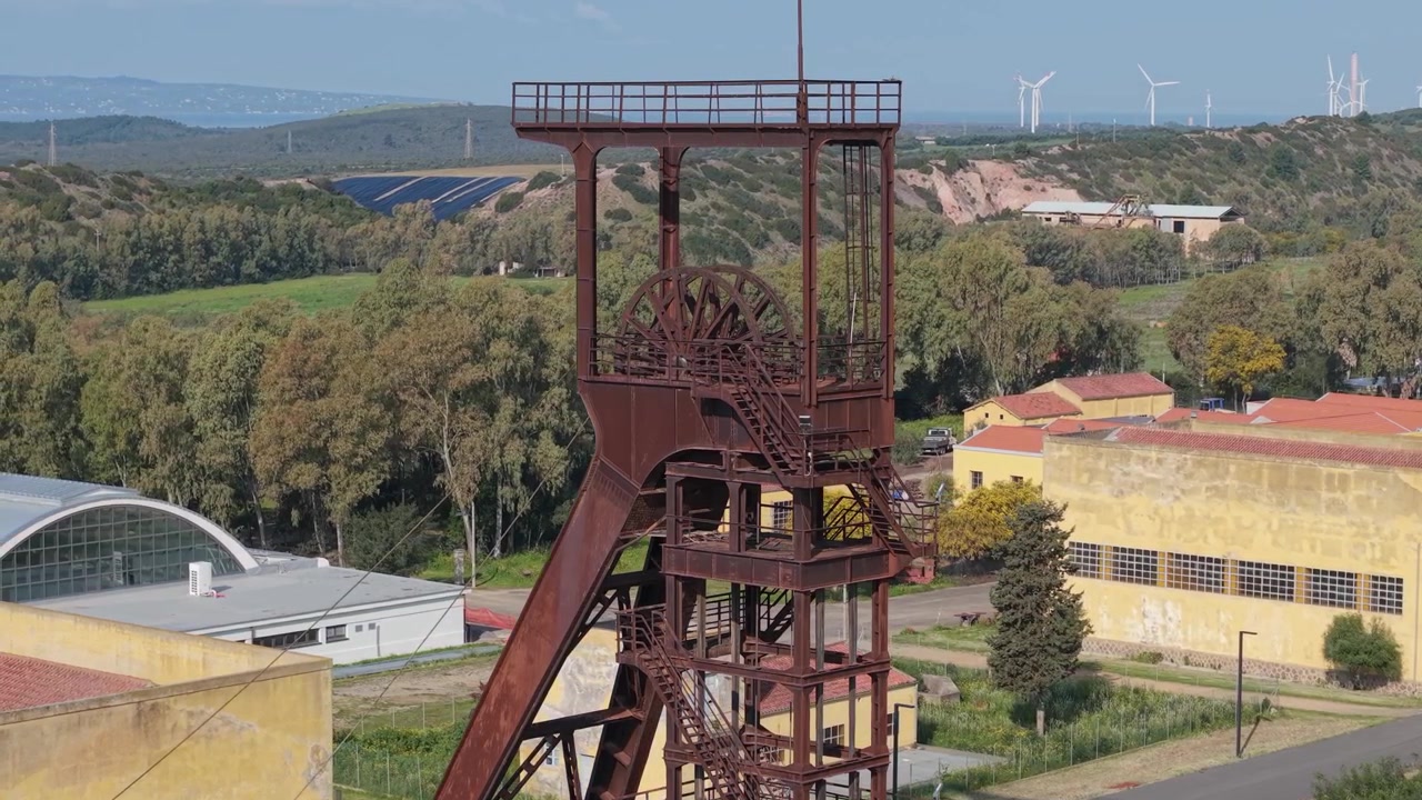 Cavaletes de mineração enferrujados na Mina de Carvão de Serbariu com uma fazenda solar e um parque eólico ao fundo em Carbonia, Sardenha, Itália. - Vista aérea