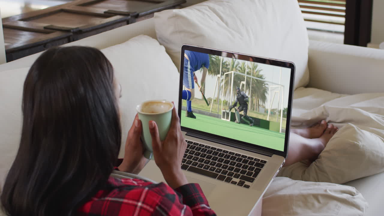 Composite of woman sitting at home holding coffee watching hockey match on laptop