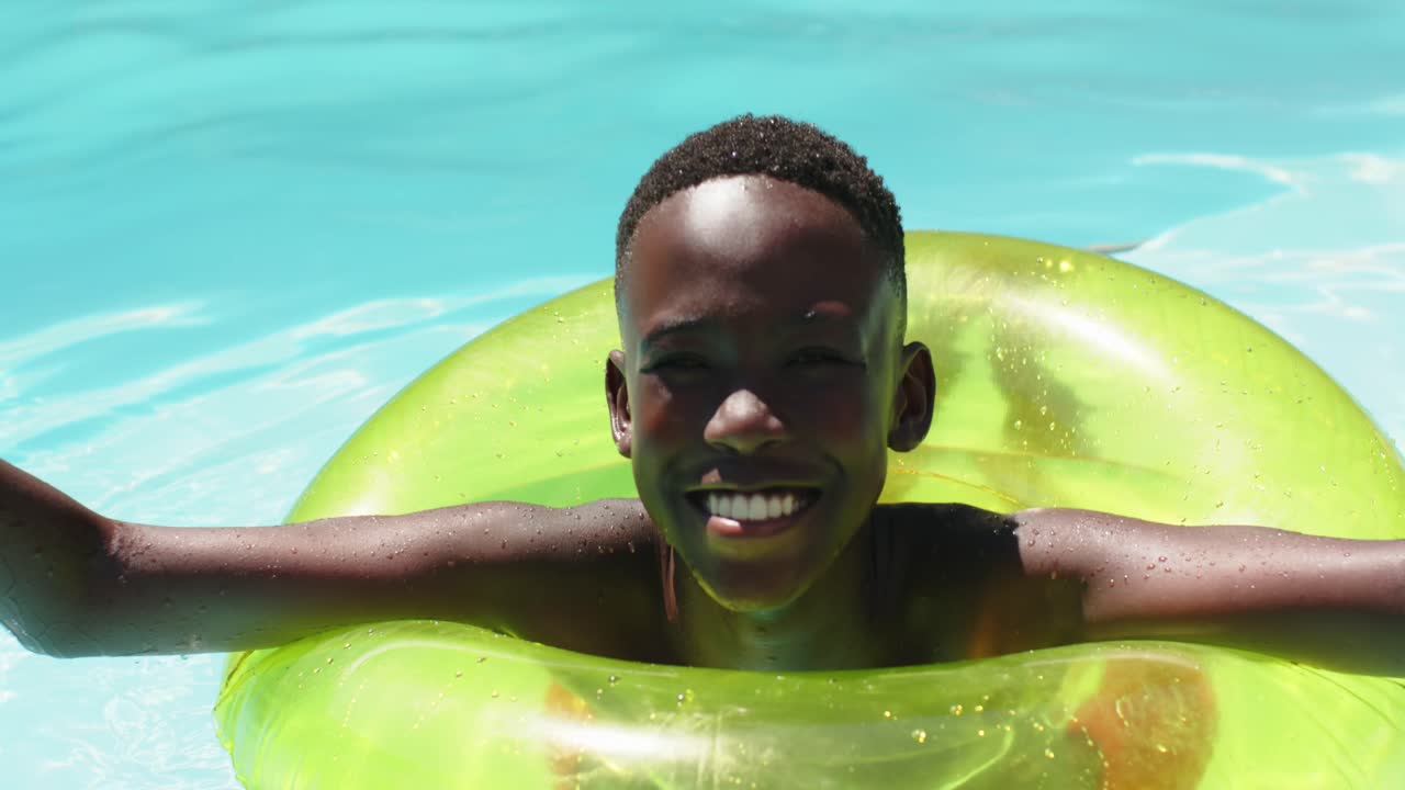 African American teen resting in neon-green float in backyard pool, seeing camera and waving