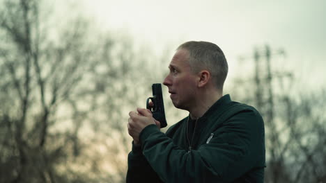 a close view of a man holding a handgun with both hands, looking around, in an outdoor setting with trees in the background