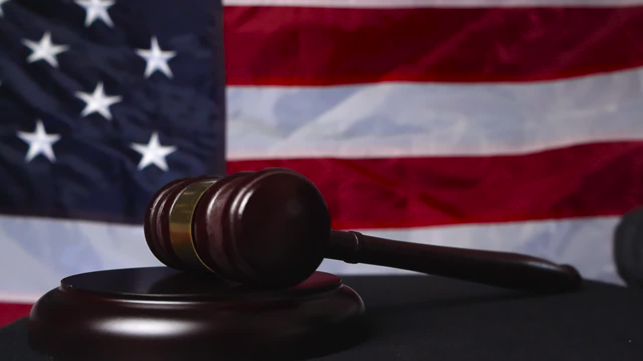 Close up view of a gloved judge justice hand hitting a court gaol wooden and bronze hammer on a black table while an American colorful United States flag is in the background. Slow motion movement