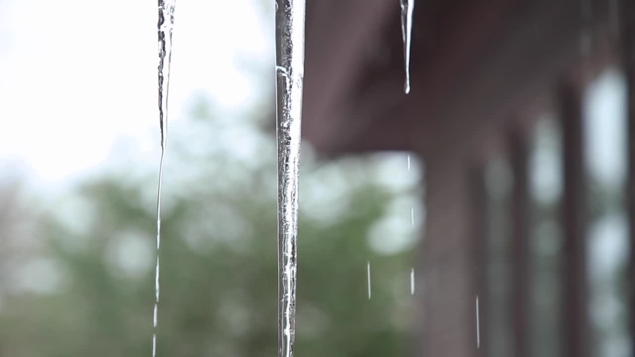 Water cascading down icicles hanging from a roof, with the side of a house and outdoor background in view.