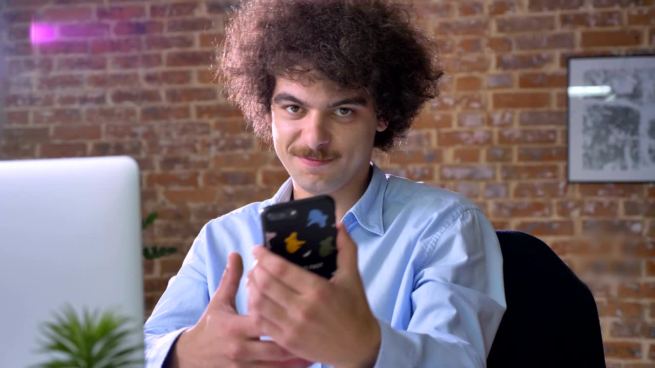 gracioso jefe nerd con volumen cabello rizado escribiendo en el teléfono y mostrando pantalla en la cámara, sentado en una oficina moderna
