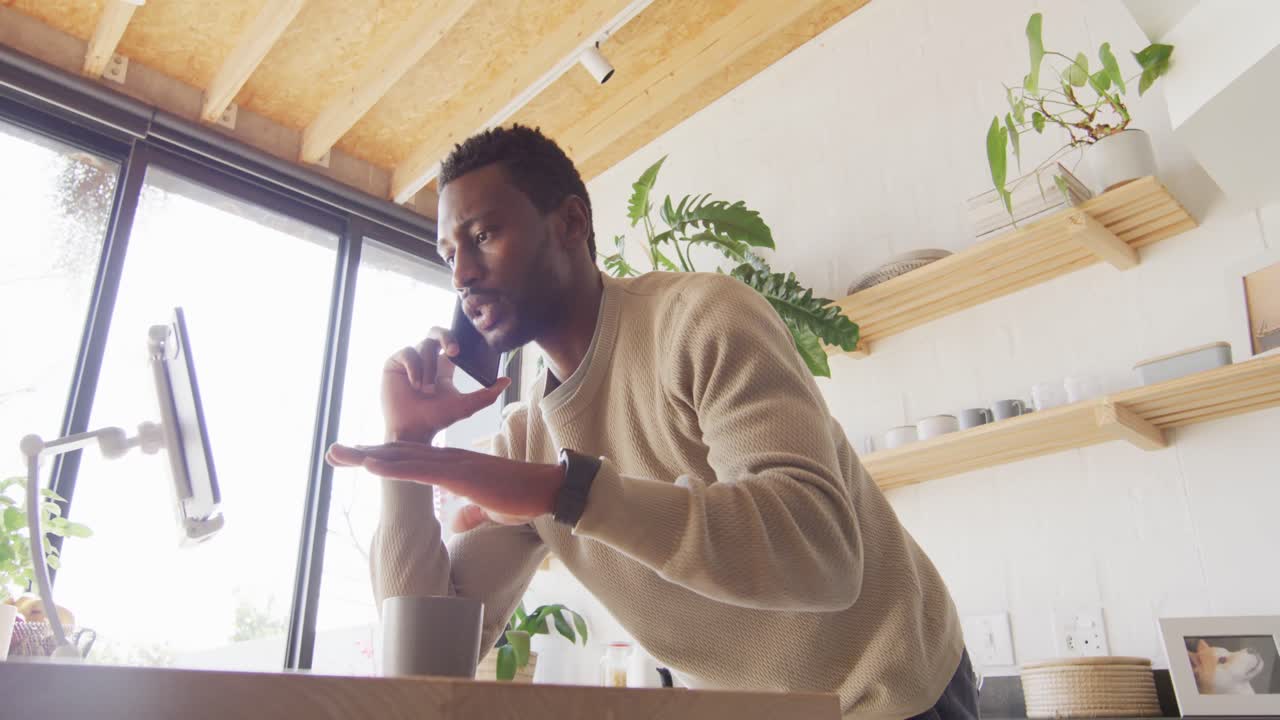 feliz hombre afroamericano apoyado en el mostrador en la cocina, usando una tableta y hablando en un teléfono inteligente