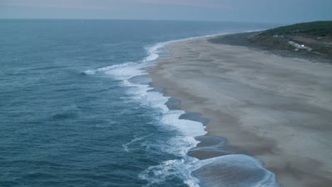 Grandes-Olas-Rompen-En-La-Costa-De-Nazare,-Portugal,-Tomadas-Desde-Arriba-Con-Vistas-Al-Océano-Y-La-Playa-Con-Algo-De-Vegetación-En-La-Distancia