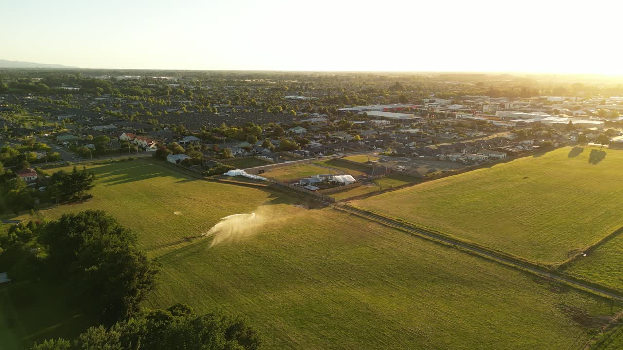 A drone flies over green New Zealand fields, capturing an irrigator in action, leading towards a distant town