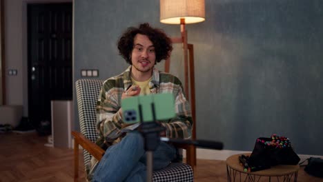un chico moreno feliz con el cabello rizado en una camisa a cuadros habla con sus suscriptores de blog usando un teléfono inteligente verde y escribe cosas importantes en casa en una habitación atmosférica