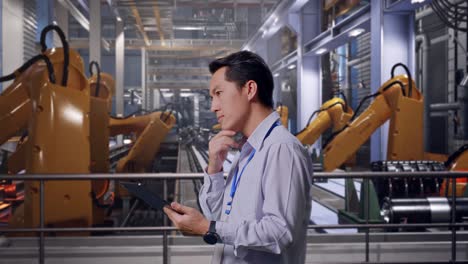 side view of an asian male professional worker with industrial robotic arms working, observes by looking up before he come to concentrating on the tablet and keep on checking