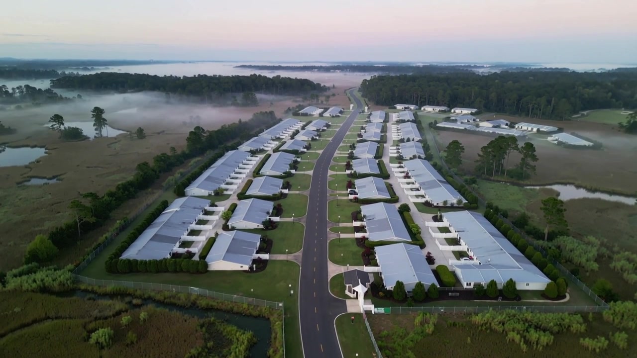 Drone che solleva la telecamera e scivola in avanti, rivelando un cancello, una strada curva e case all'alba, con una palude