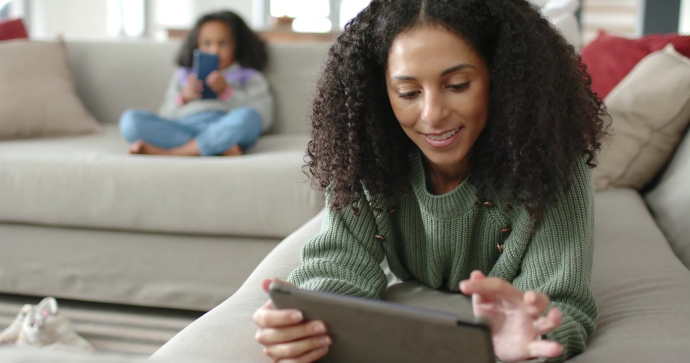 African mother tapping tablet while camera shifting focus, daughter on sofa scrolling smartphone