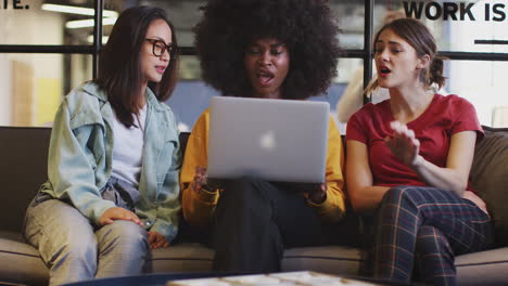 Three-millennial-women-sitting-on-the-sofa-in-an-office-lounge-using-a-laptop-together,-tilt-shot