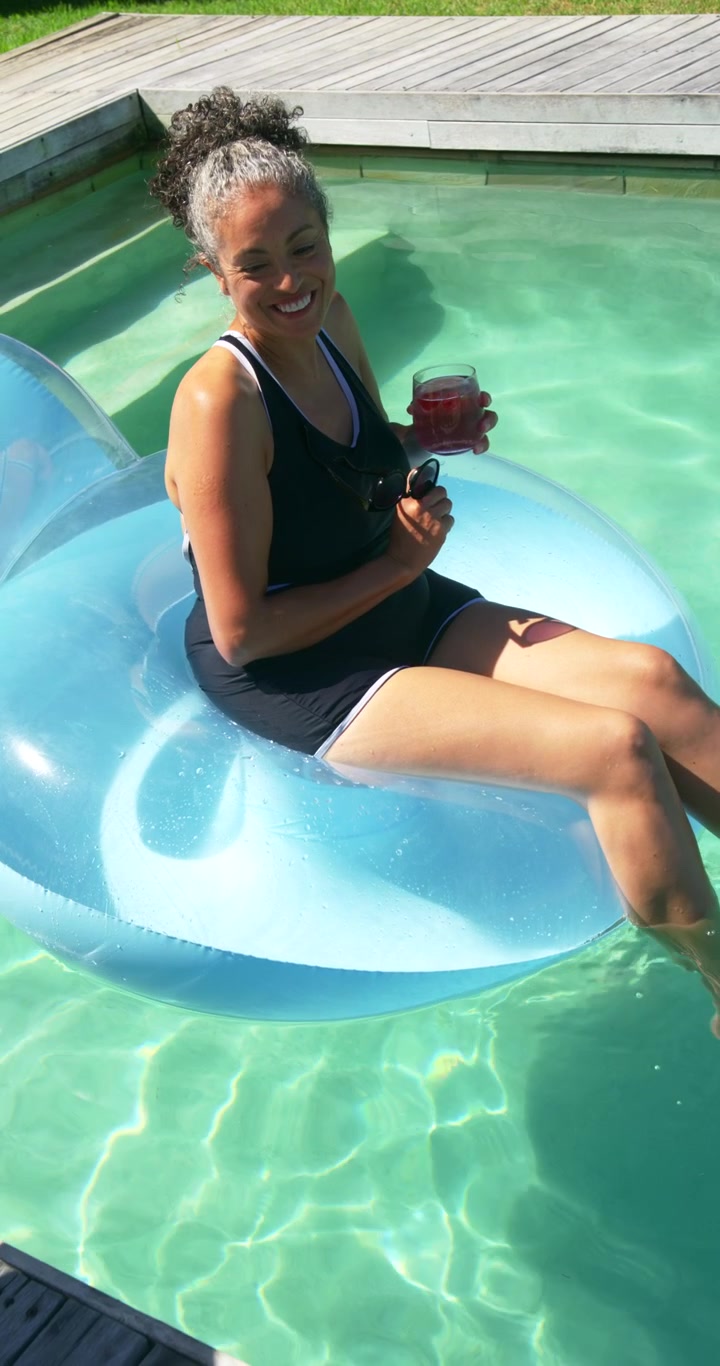 Vertical video: African American woman sitting on float in pool holding drink, relaxing during pan