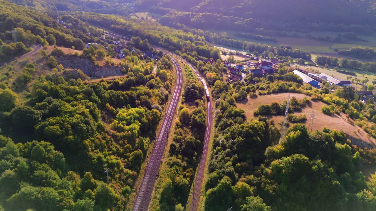 Drone view of passenger train moving along winding railway through forested hills and farmland towards rural village