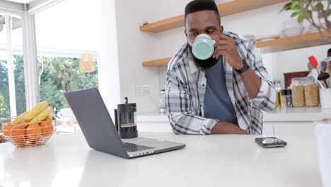 african american man making coffee and using laptop in sunny kitchen, slow motion