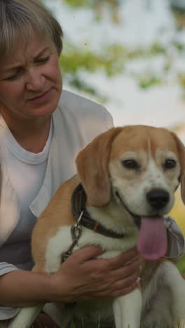 mujer sentada en un campo cubierto de hierba sosteniendo a su perro con afecto mientras acaricia suavemente al perro, ambos disfrutando de un momento pacífico al aire libre, el fondo presenta vegetación exuberante y árboles bajo la cálida luz del sol