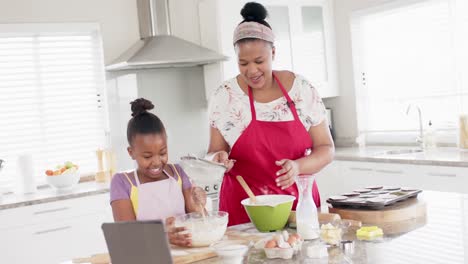 Happy-unaltered-african-american-mother-and-daughter-baking-in-kitchen-using-tablet,-in-slow-motion