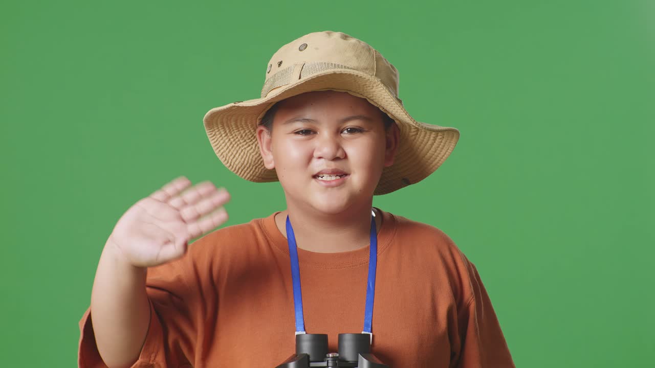 모자와 안경을 쓴 아시아 관광 소년, 초록색 스크린 배경에 서 있는 동안 미소 짓고 손을 흔들며, 소년 연구자는 무언가를 조사합니다, 여행 모험, 클로즈업