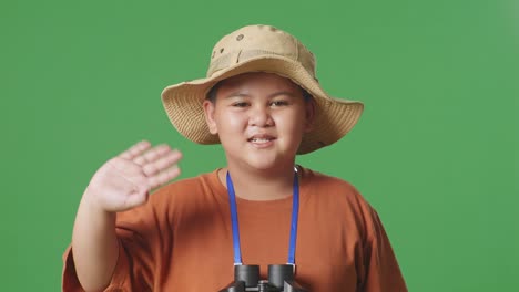 asian tourist boy with hat and binoculars using magnifying glass, smiling and waving hand while standing on the green screen background. boy researcher examines something, travel adventure, close up